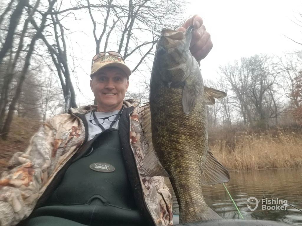 A man in a baseball cap and full fishing gear holding up a Smallmouth Bass caught in Lake Hopatcong against a background of bare winter foliage on a cloudy day