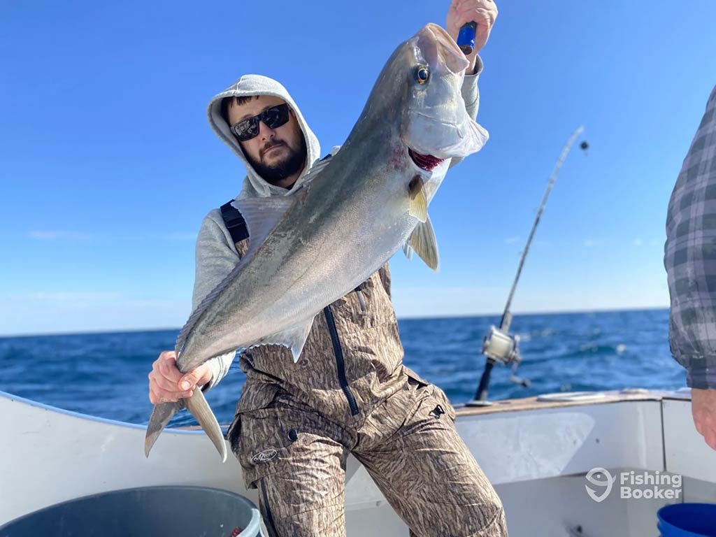 A man in a hoodie and sunglasses standing on a deep sea fishing charter on the open waters of the ocean and holding a large Tilefish on a clear day