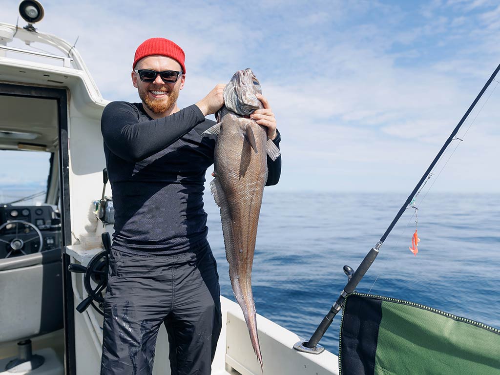 An angler in a red hat and sunglasses standing on a fishing charter on the deep, open waters and holding a large Grenadier fish with both hands, as a rid leans on the side of the boat next to him