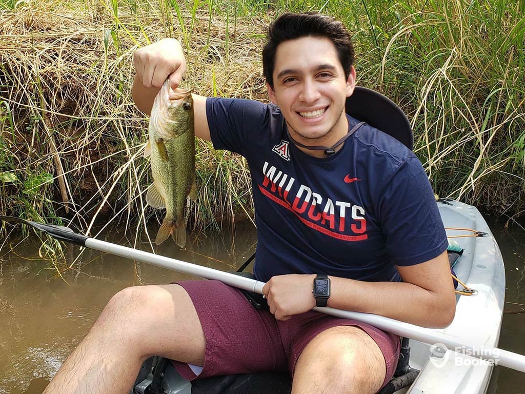 A young man sitting in a kayak and smiling as he holds a small Largemouth Bass in one hand and his paddle in the other
