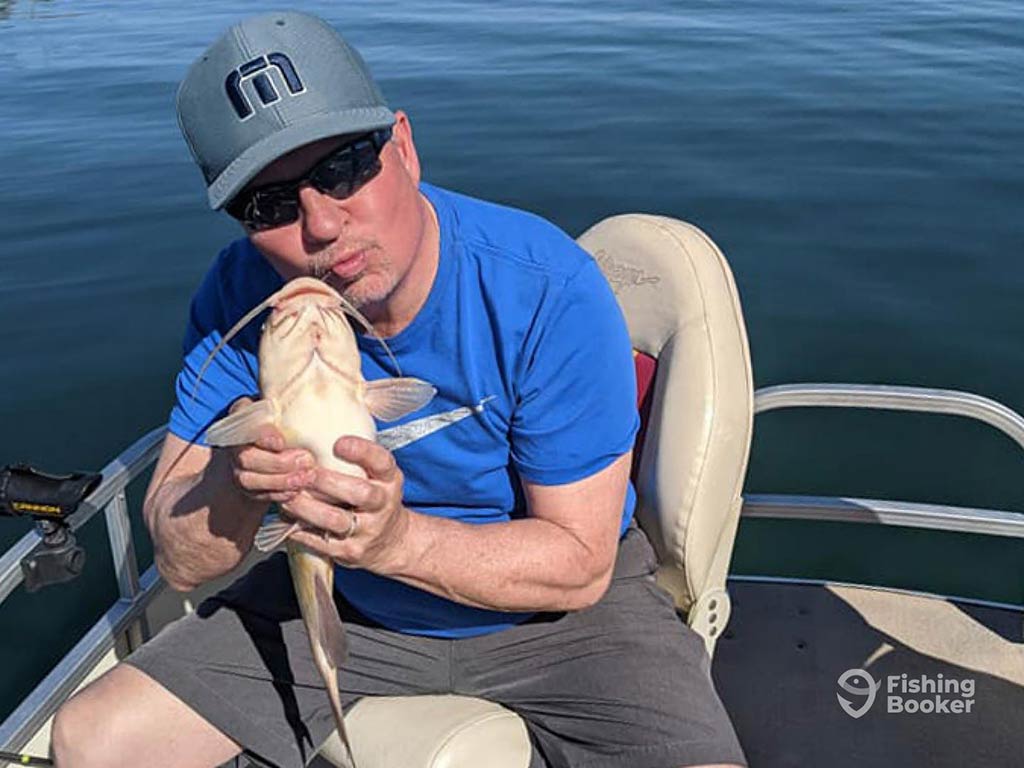 A man in a baseball cap and sunglasses sitting on a fishing boat and holding a small Catfish as he pretends to kiss it on a sunny day