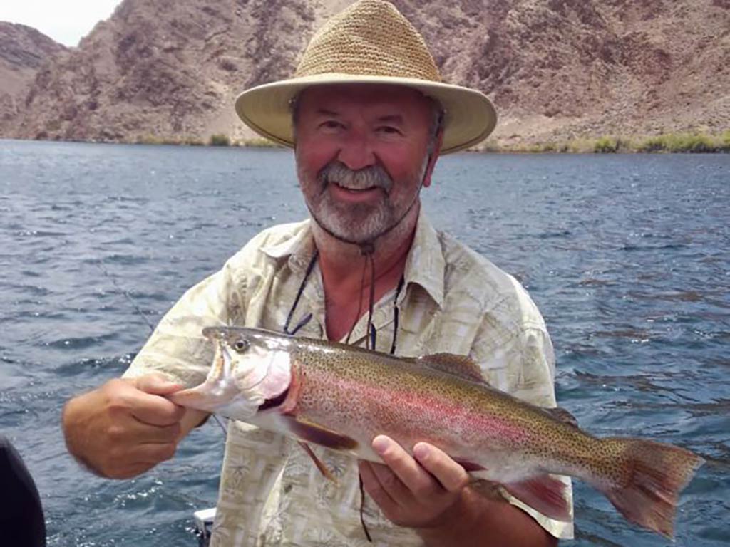 A man in a straw har smiling on a fishing boat as he holds a Rainbow Trout against a background of a river or lake and dry, arid mountains