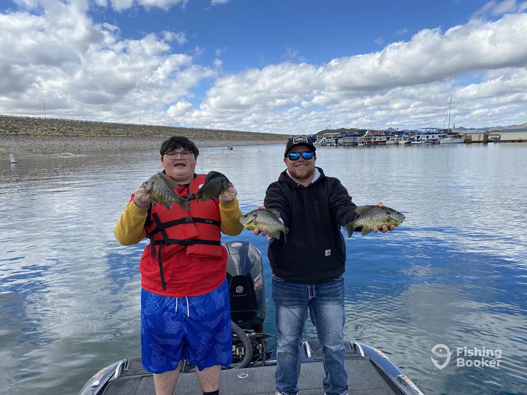 Two young men aboard a fishing boat on calm waters in Arizona on a day with sunny intervals, with both holding two Bluegill fish each