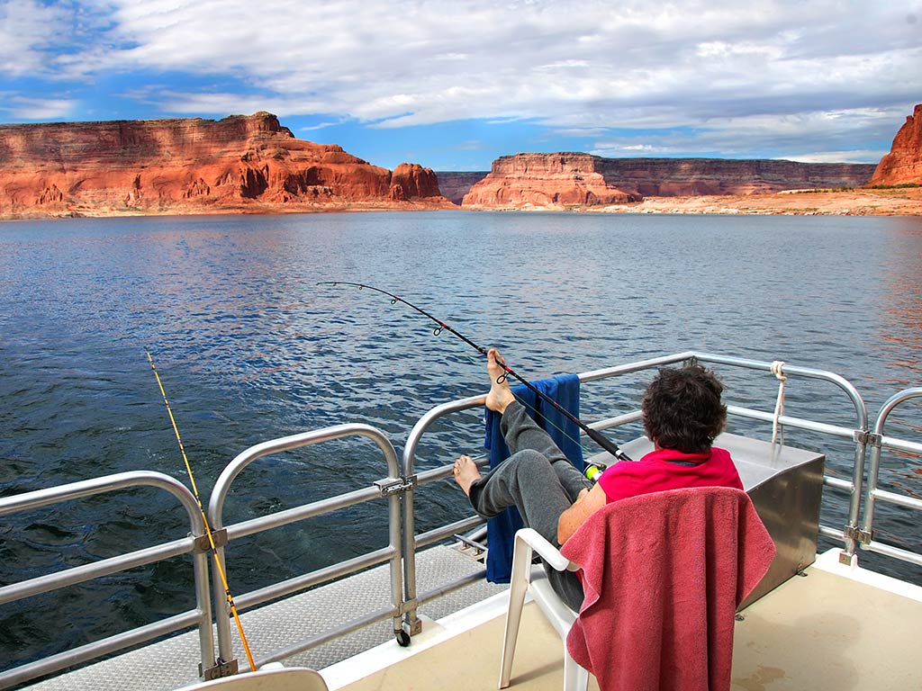 A view from behind of a woman sitting on a boat and fishing with her legs up and her rod resting between her toes on a lake looking towards impressive rock formations in the distance