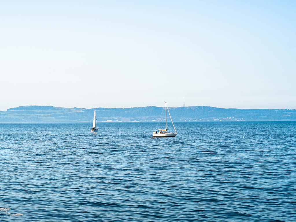 Sailboats on the calm waters of the North Sea with distant hills behind Edinburgh