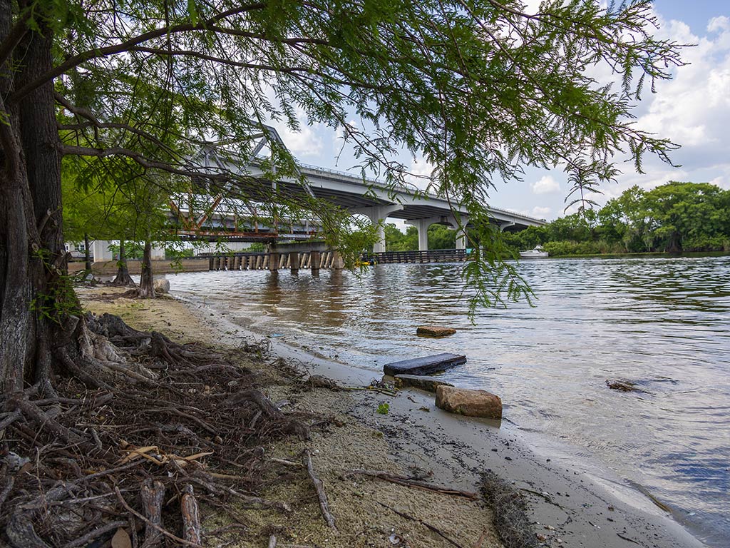 A calm river flows along a sandy, tree-lined shore, perfect for fishing in Lake Monroe. A concrete bridge crosses the water and is supported by sturdy pillars. Debris like rocks and logs is scattered along the shoreline, while the sky's partly cloudy.