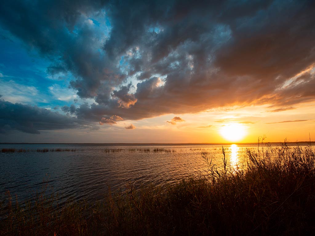 A sunset over a calm body of water paints the sky with a gradient from blue to orange, and dramatic clouds partially cover the horizon. Silhouetted grass and reeds grace the shoreline, hinting at a tranquil evening.