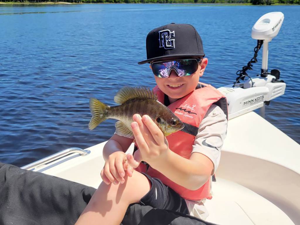 A child wearing a baseball cap, sunglasses, and a life jacket proudly holds a small Fish while sitting in a boat on Lake Monroe. The gentle ripples of the water, and part of the boat's motor create a perfect backdrop for this fishing adventure.