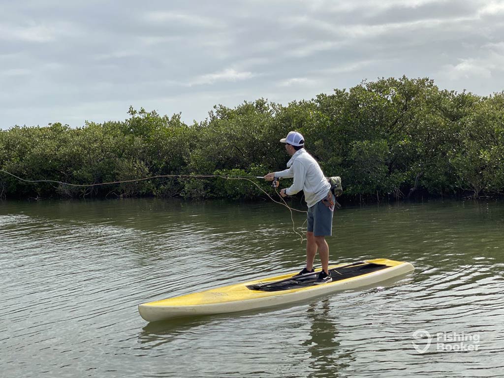 A person wearing a cap stands on a yellow paddleboard in the calm waters of Lake Monroe, holding a fly fishing rod. Dense green Shrubs line the water's edge in the background, as cloudy skies loom overhead.