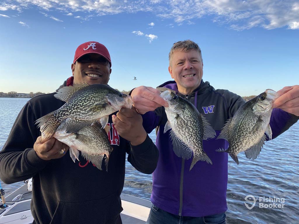 Two men stand on a boat, smiling and holding the Crappie they caught while fishing in Lake Monroe. The man on the left is wearing a red cap and black jacket, while the one on the right is sporting a purple jacket. The sky is blue with scattered clouds, and a bird can be seen in the background.