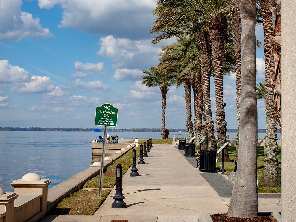 A paved walkway, lined with palm trees, stretches alongside a calm body of water under a partly cloudy sky. A sign reads, "No Skateboarding or Skating on Walk." In the distance, a few people are near a seating area.