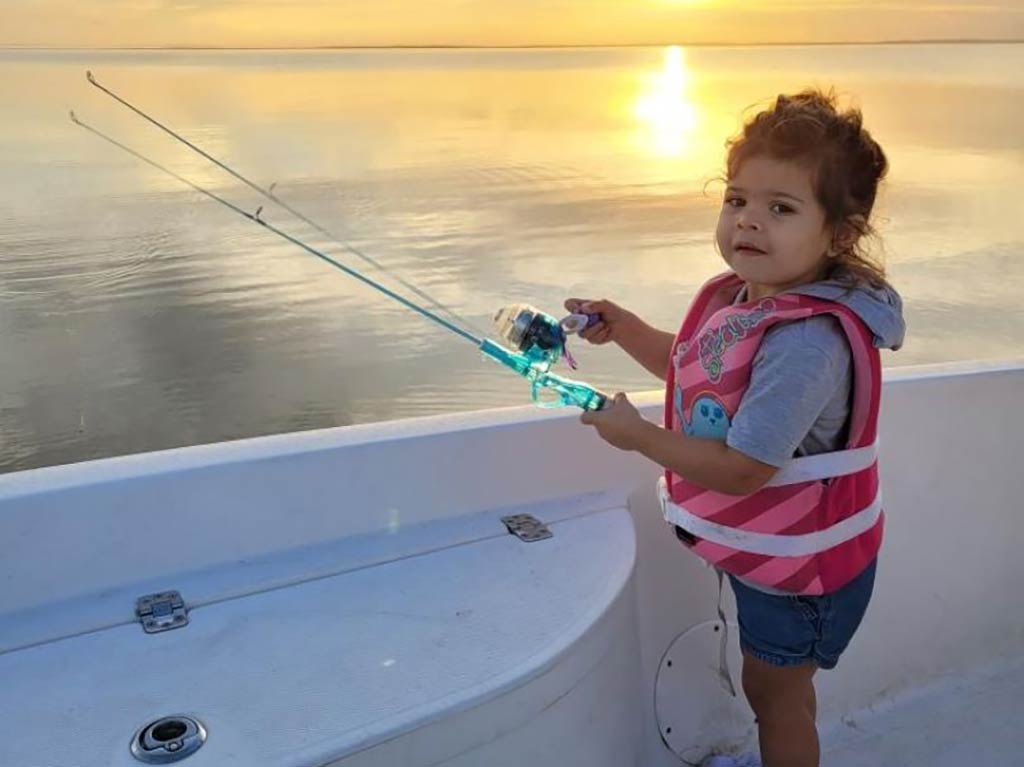 A young child wearing a pink life vest and denim shorts holds a fishing rod on a boat. In the background, there's a calm body of water, and the sky is painted with a golden sunset.