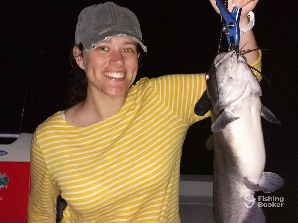 A woman in a yellow and white striped shirt and a cap is smiling while holding a large Catfish, with a blue tool at nighttime.