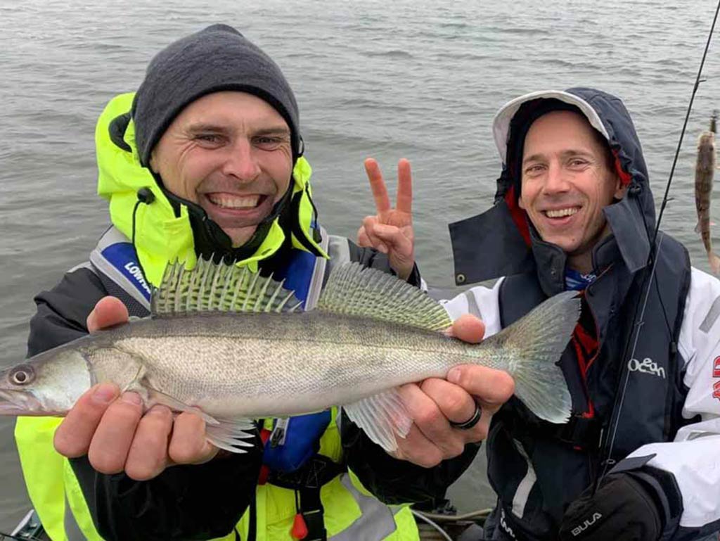 Two people, dressed in winter jackets, on a boat holding a Zander fish. One person smiles and raises the fish with both hands, while the other makes a peace sign and smiles