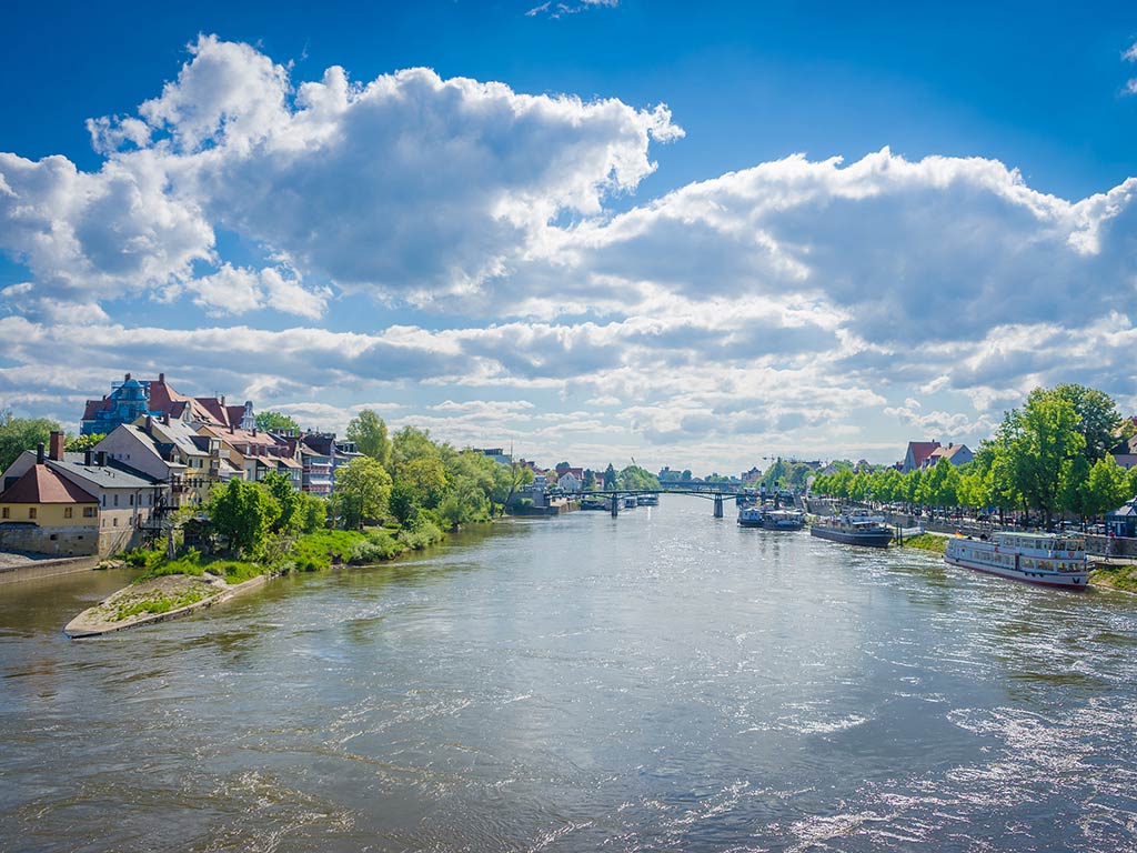 The Danube River flows between two banks, lined with trees and buildings, under a blue sky with scattered clouds. Boats are anchored along the shores, and a distant bridge spans the waterway