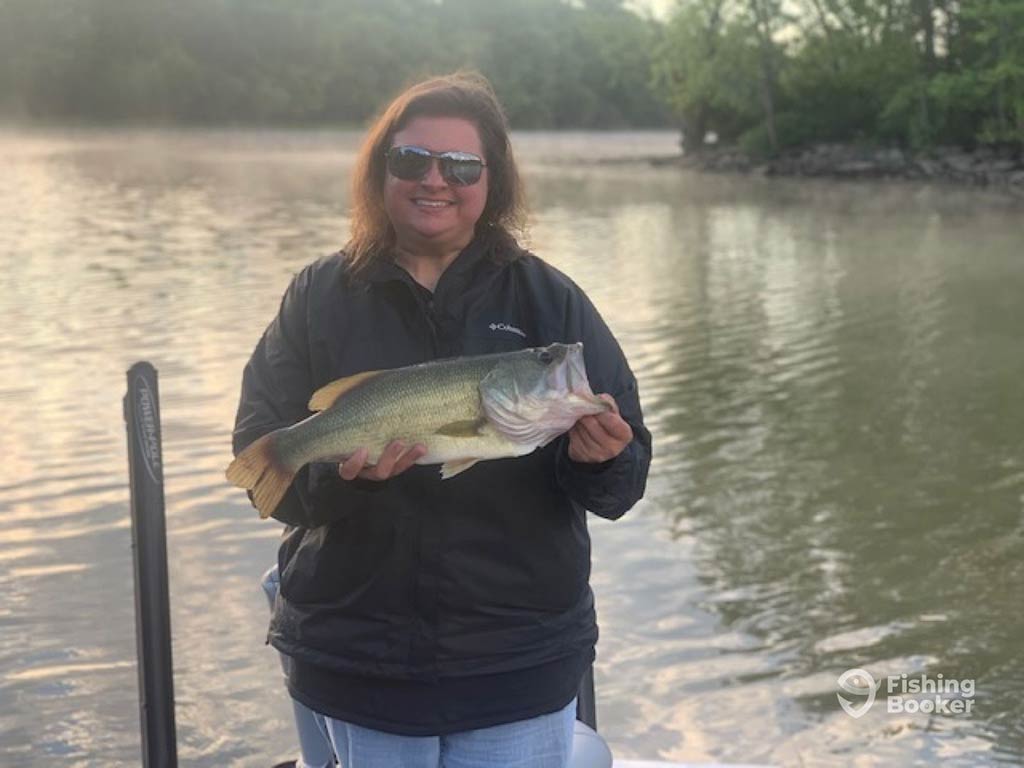 A woman standing on a dock and smiling as she holds a Largemouth Bass after a successful fishing trip in New England 