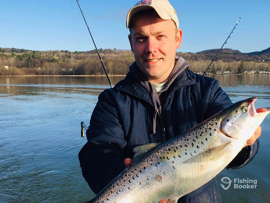 A man in a baseball cap smiling as he poses with his Landlocked Salmon catch next to a calm lake in New England on a sunny day