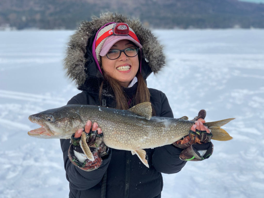 A girl in winter clothes standing on a frozen lake holds a Lake Trout that she caught while ice fishing somewhere in the US.
