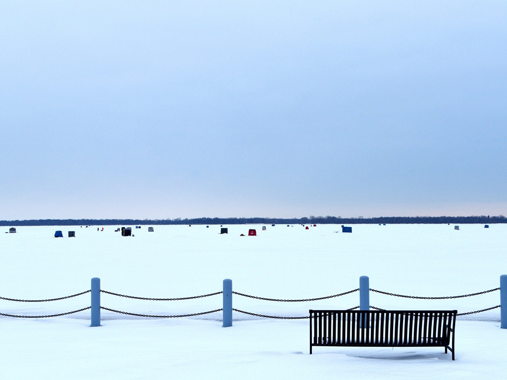 A snow-covered landscape with a bench in the foreground. Several small, colorful structures dot the snow in the distance under a cloudy sky.