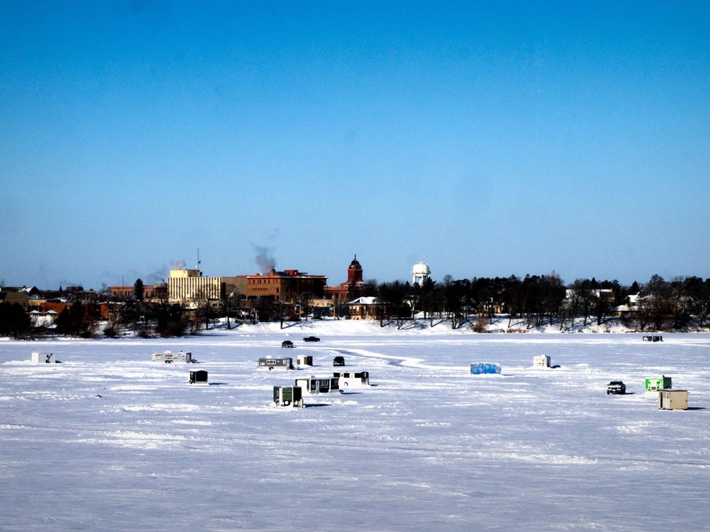 A frozen lake with ice fishing huts scattered across its surface, city buildings visible in the background under a clear blue sky.