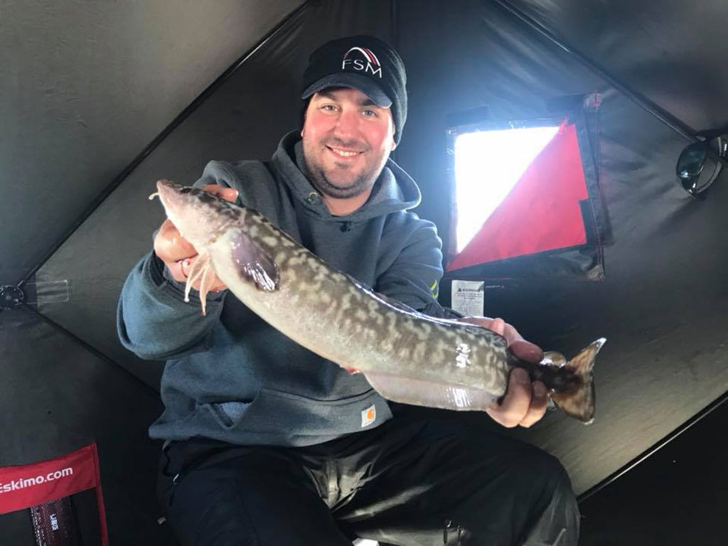 A man in winter clothing's holding a sizeable Burbot inside an ice fishing tent.