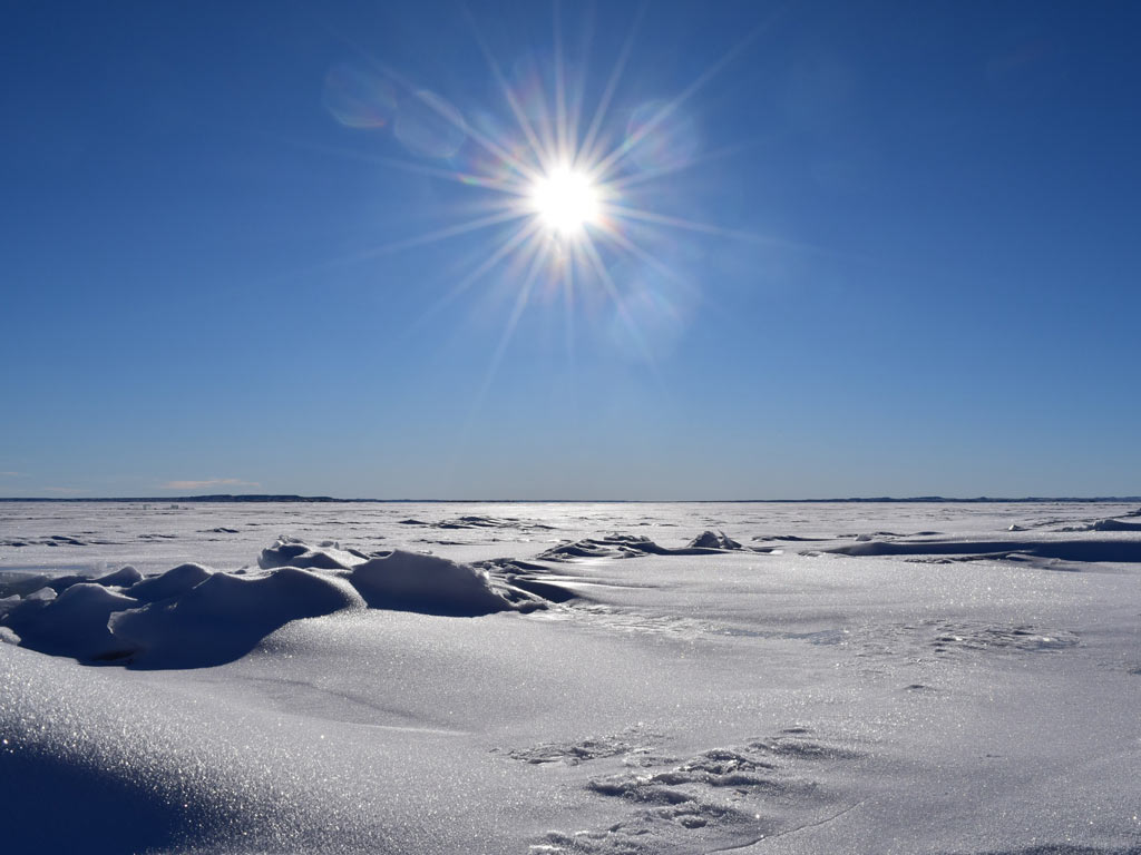 A bright sun is shining over a vast, snowy landscape of a frozen Fort Peck Lake, with clear blue skies above.