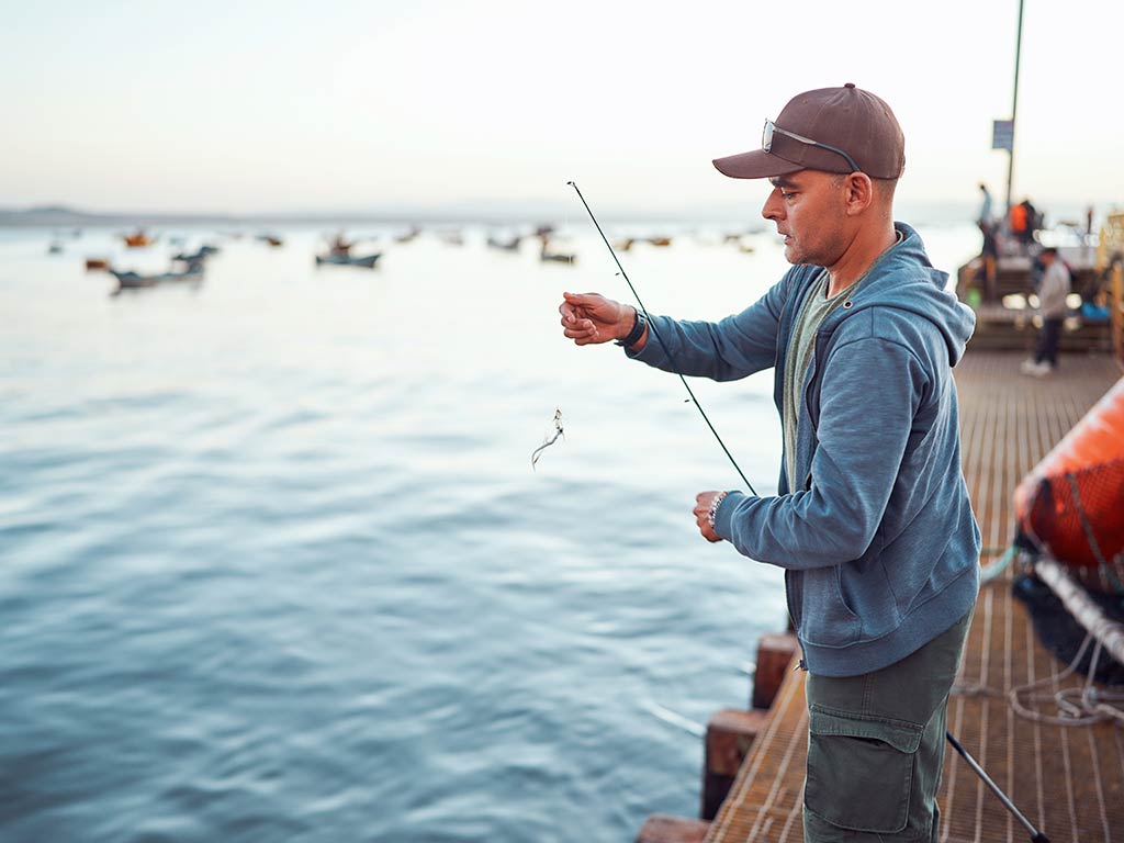 A man standing on a dock, holding a fishing rod with a small fish. In the background, you can see boats on the water.