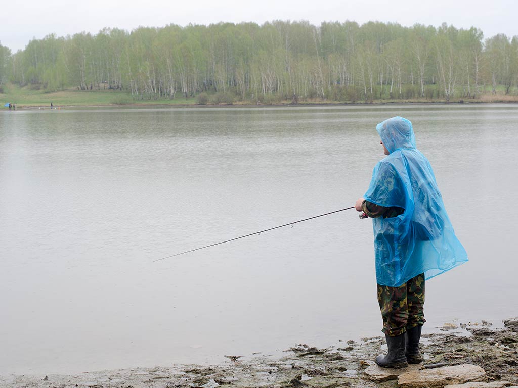 A person in a blue rain poncho enjoying Bass fishing by the lake, while the forested shoreline provides a serene backdrop