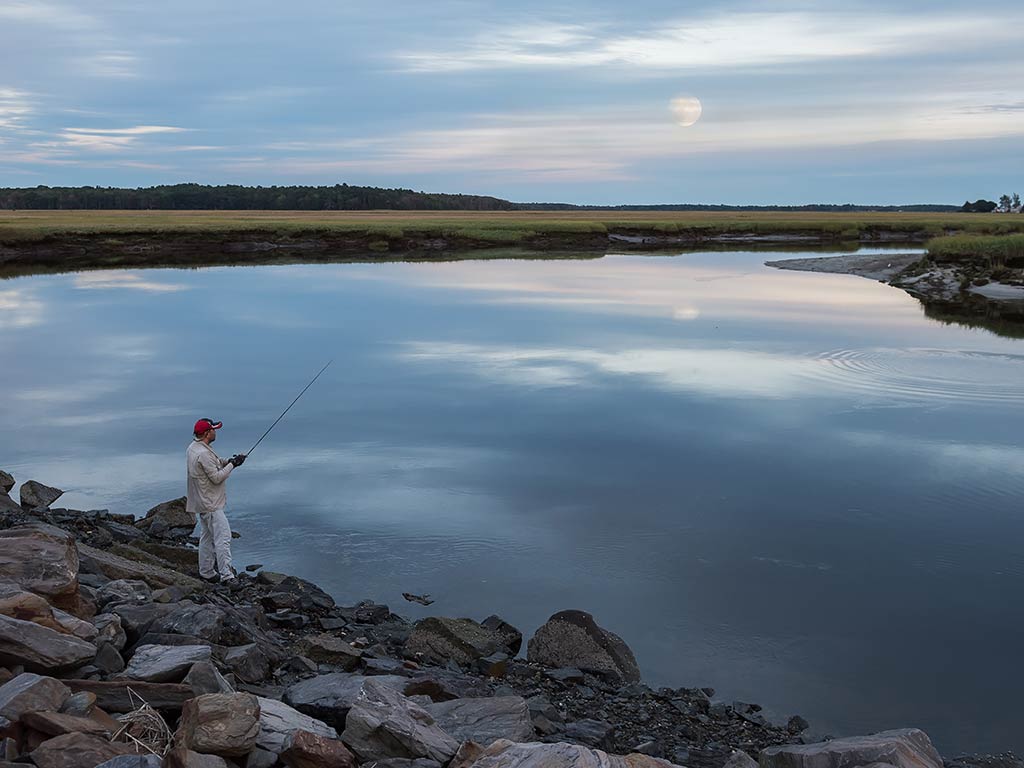 A person enjoys fishing by a calm river under a cloudy sky, with a distant full moon on the horizon. Rocks and grassy banks surround the tranquil waters