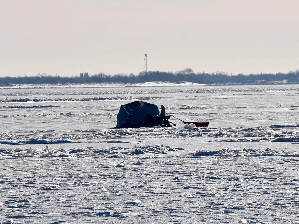 A distant ice fishing tent on a vast frozen Lake Winnipeg under a clear sky, with an ATV and sled set up nearby for the perfect ice fishing adventure.