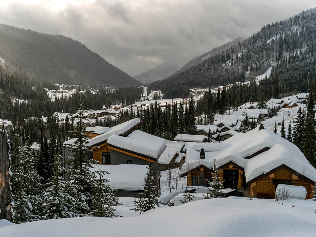 Snow-covered cabins nestled in the mountainous valley in the town of Kamloops surrounded by dense pine forests under a cloudy sky.