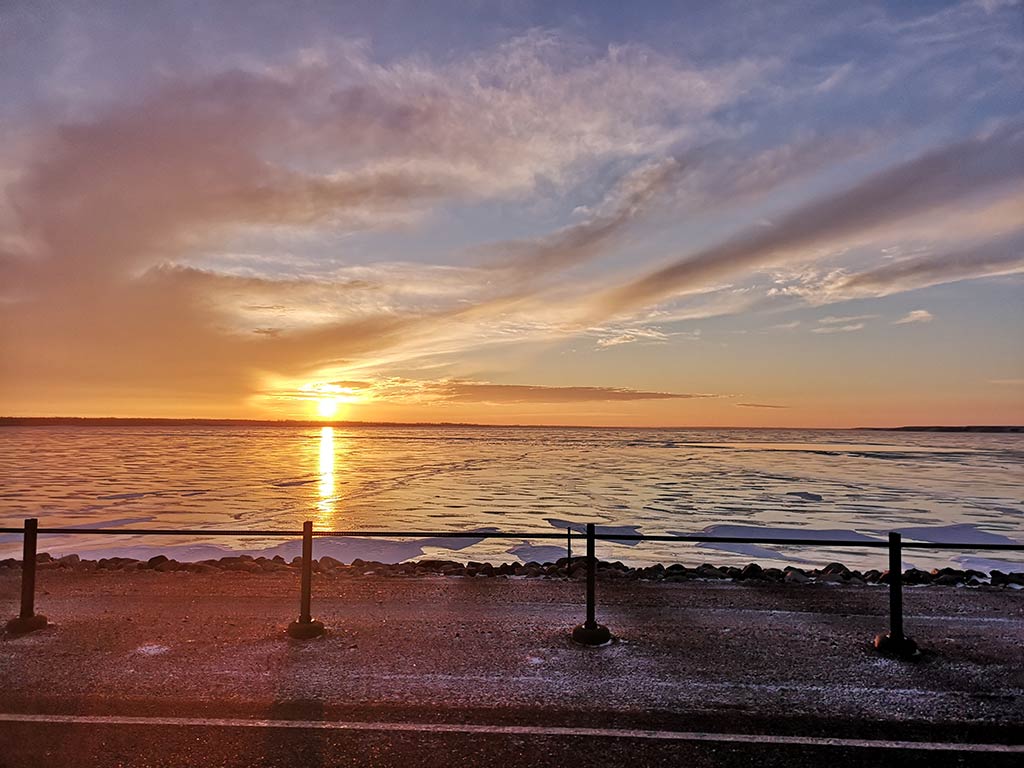 Sunset over a frozen lake, Diefenbaker with clouds scattered across the sky. A barrier fence lines the foreground, hinting at ice fishing as the icy surface reflects the orange hues of the setting sun.