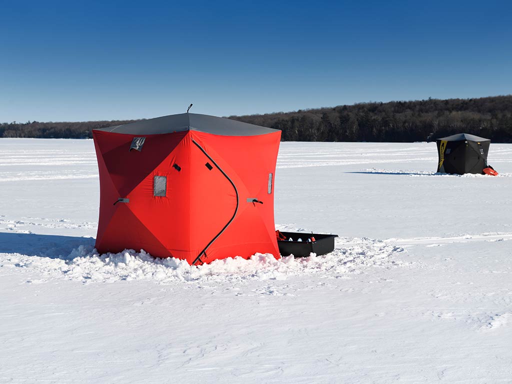 Two tents for ice fishing sit on a frozen lake in Canada under a clear blue sky. The closer tent is red, while the distant one is darker, both surrounded by pristine snow.
