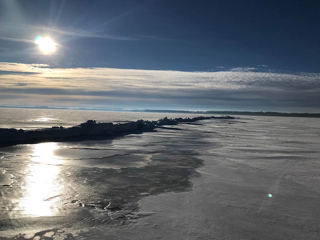 A frozen landscape of Gull Lake in Canada with cracked ice glistens under a bright sun, reflecting on the icy surface.