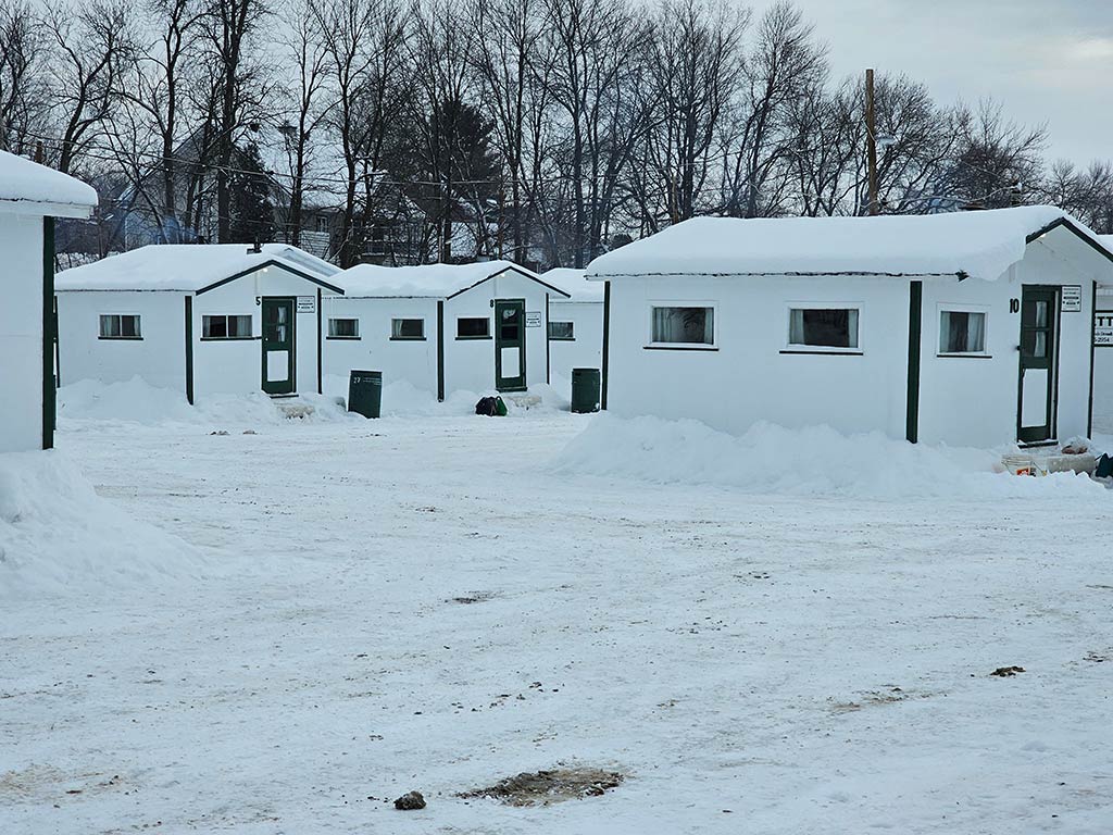 Small white cabins in a snowy area of Trois Rivieres in Canada, perfect for ice fishing, are surrounded by bare trees. 