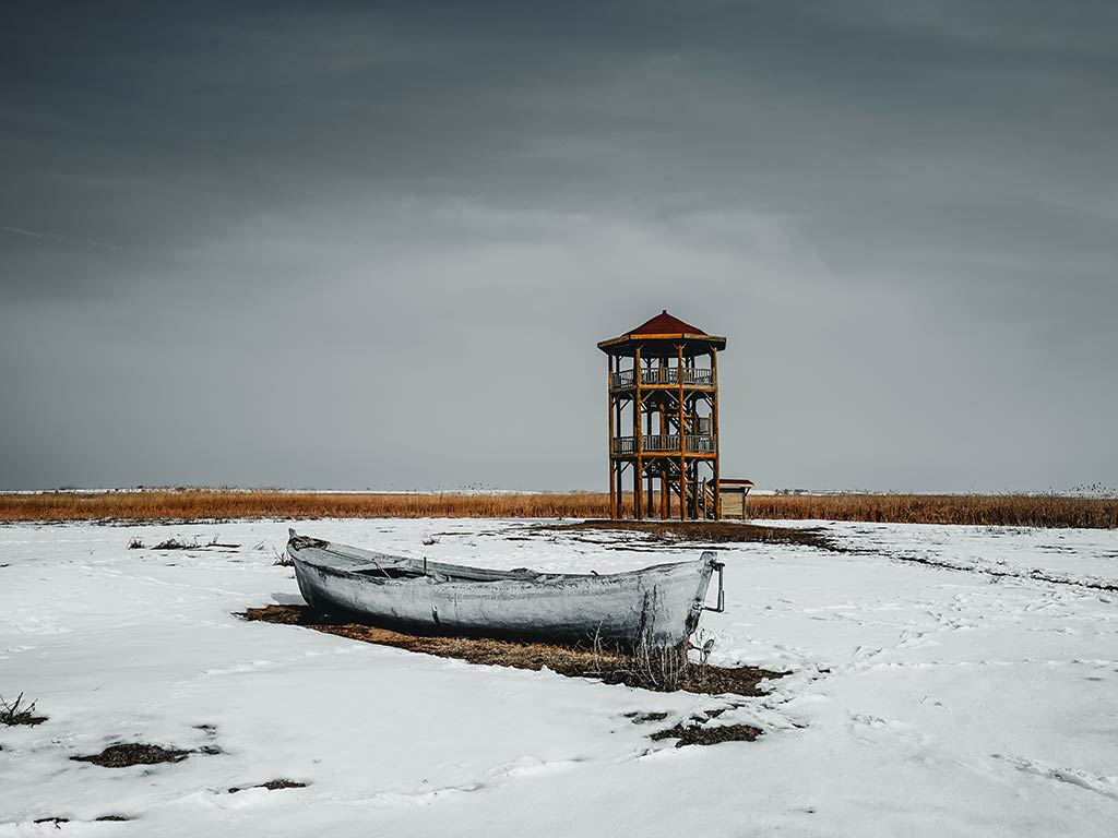 A small, weathered boat rests on a snow-covered field, evoking scenes of Canada. In the background, a wooden observation tower stands under a cloudy sky.