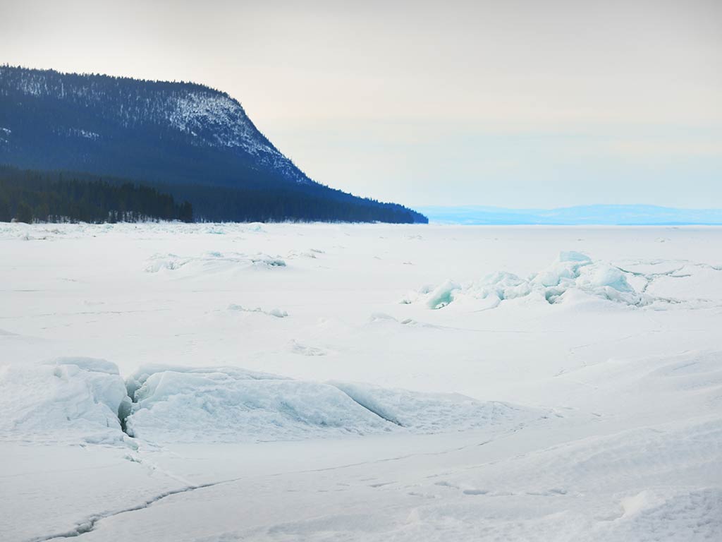 A snow-covered landscape with ice formations in the foreground, a forested hill on the left, and an overcast sky creating a picturesque scene.