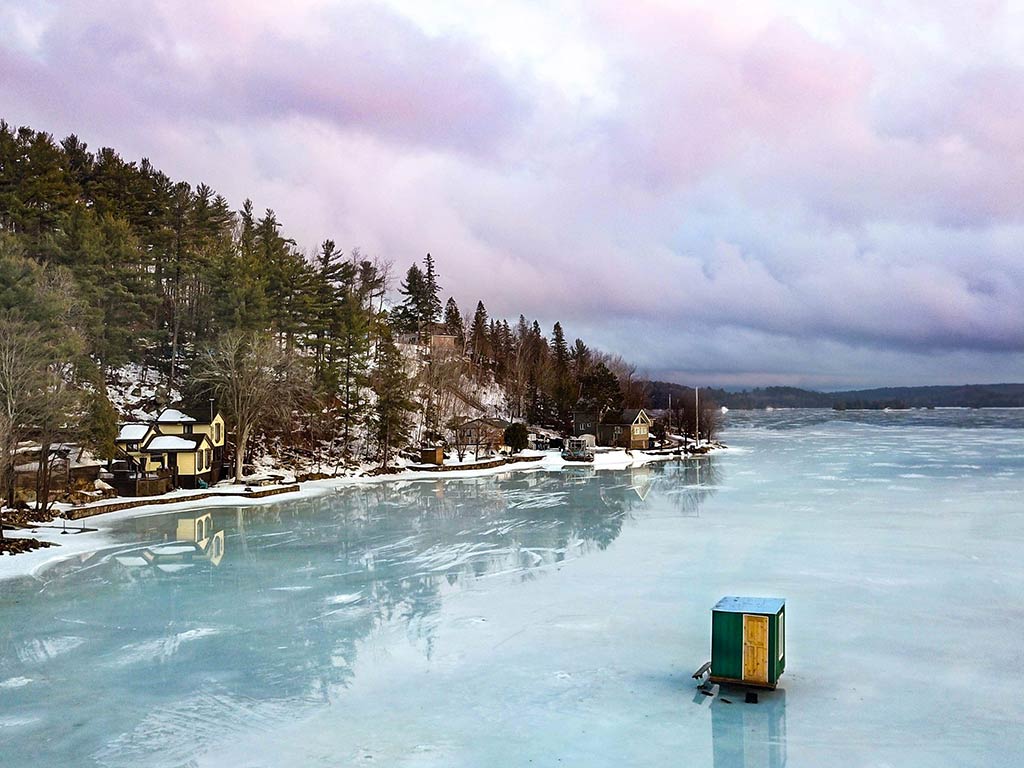 A serene, frozen lake in Canada with a small ice fishing hut visible in the foreground. Snow-dusted trees and houses line the shore under a cloudy, colorful sky.
