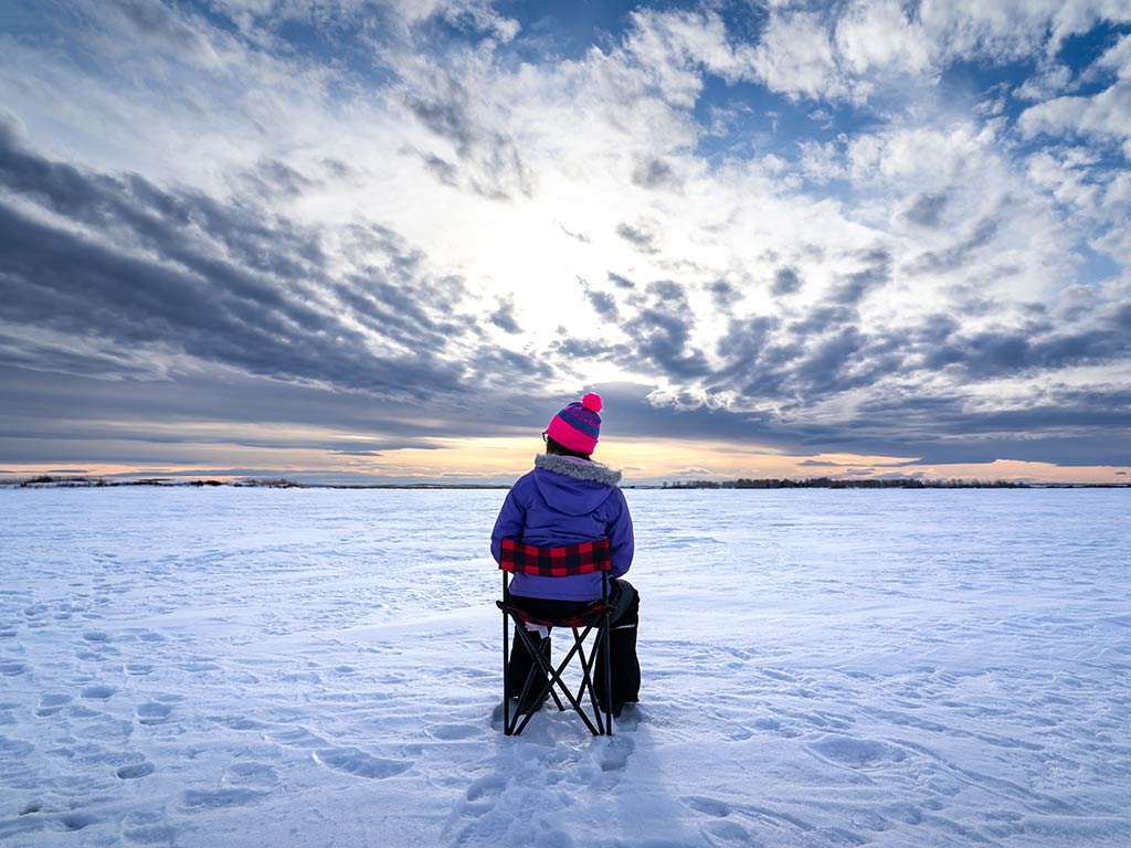 A view from behind of a  person in a purple jacket and pink hat sitting on a folding chair, ice fishing in Canada's snowy landscape under a cloudy sky.