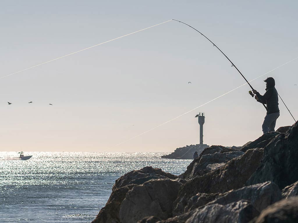 A person fishing from a rocky shore while a boat floats in the distance on a calm sea under a clear sky. In the background, there's a tall structure visible.