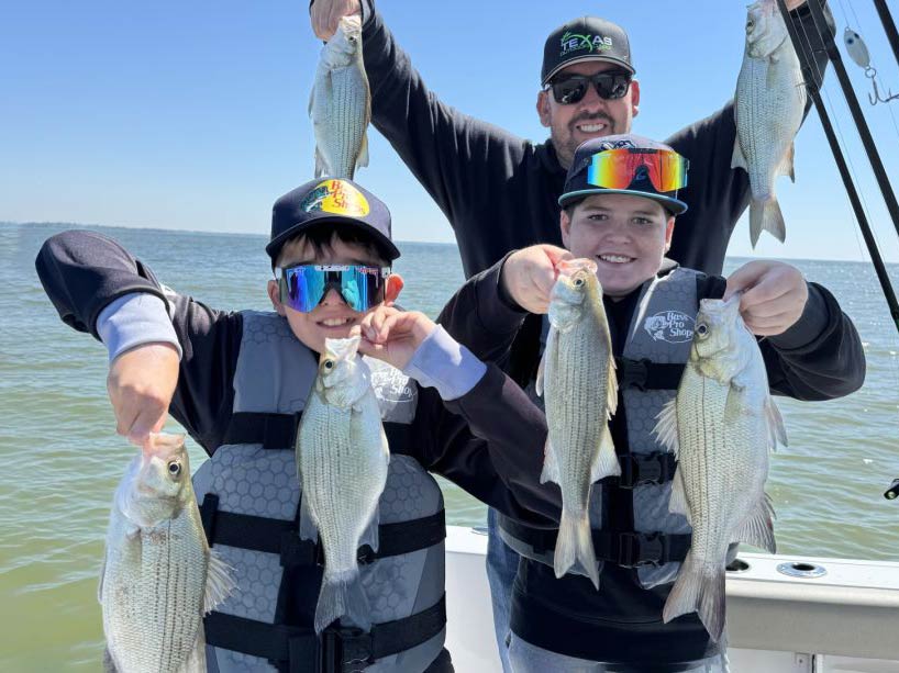 A man standing behind two children on a fishing boat on a clear day, with each of them holding two White Bass