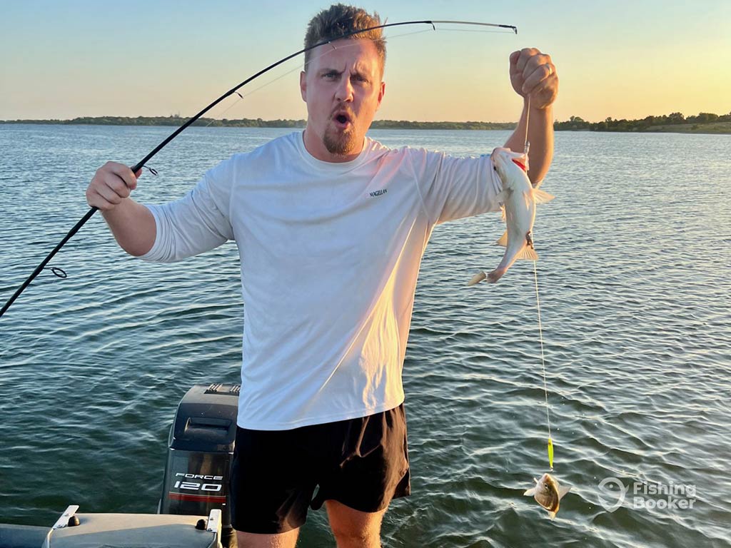 A man standing on a fishing charter on a boat and posing as he holds his fishing rod in one hand line in the other, with two White Bass on the end of it