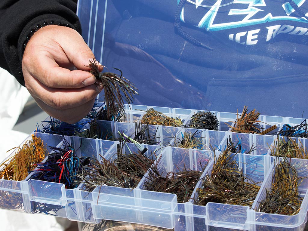 A closeup of someone holding a skirted fishing lure next to a tackle box filled with various skirt-style lures