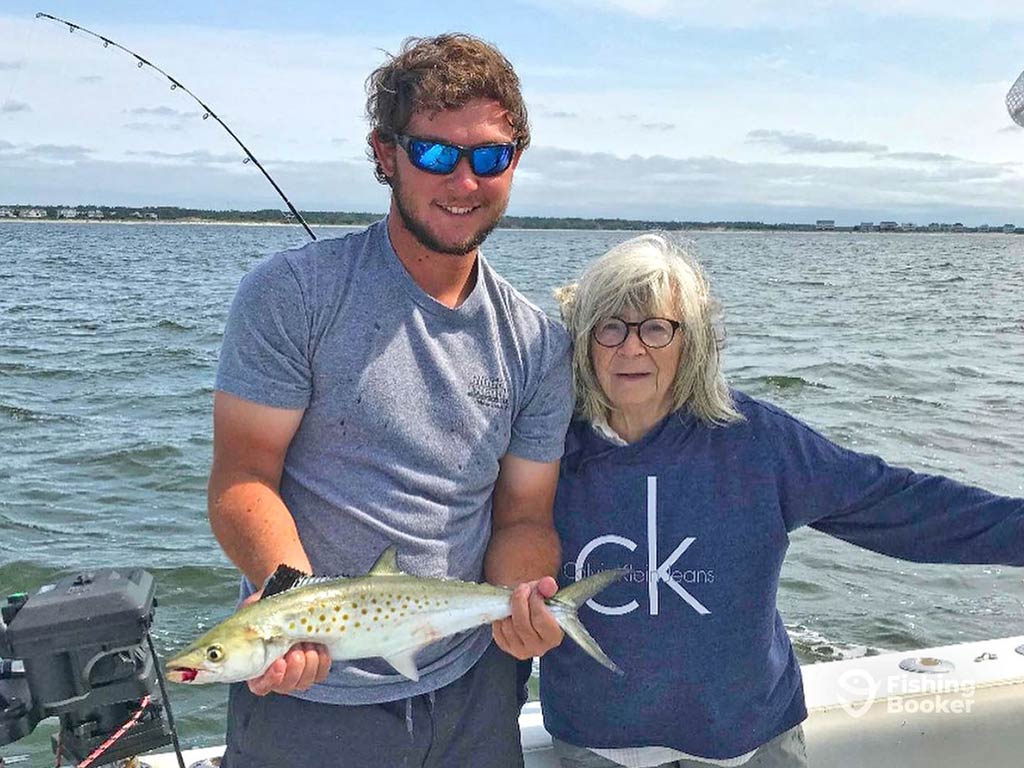 A man and an older woman stood on a fishing charter on a clear day, as the man holds a Spanish Mackerel in both hands