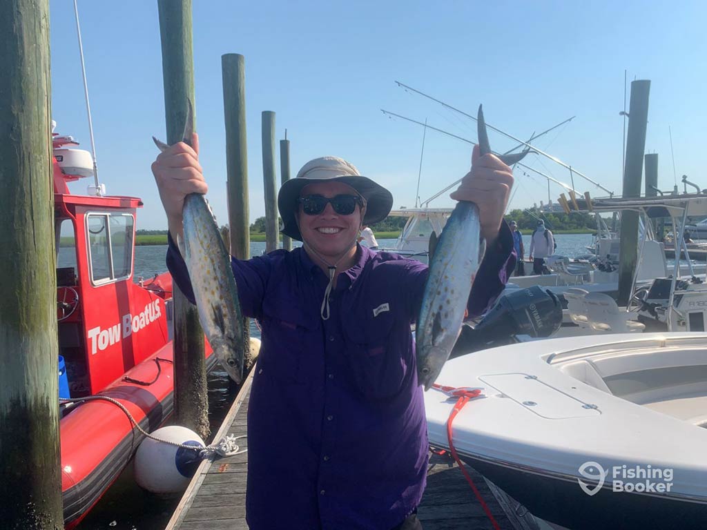 A man standing on a dock in North Carolina holding two Spanish Mackerel after a successful day's fishing, with boats visible behind him on a clear day