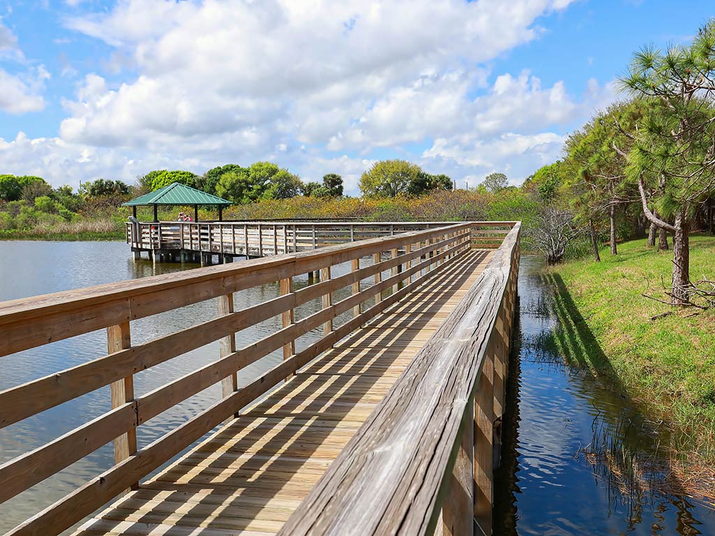 A wooden boardwalk over water leads to a covered gazebo in a park in Wakodahatchee, FL. Trees and grass are on the right, and the sky is partly cloudy.