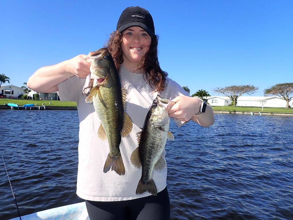 A woman holding two Largemouth Bass on a boat in a sunny lake setting, wearing a black cap and an off-white shirt.