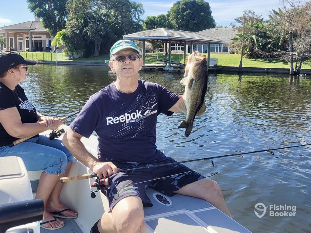 A man’s holding a fish on a boat, wearing a Reebok shirt and cap. A woman’s seated nearby with a fishing rod. The water's calm, and there are houses in the background.