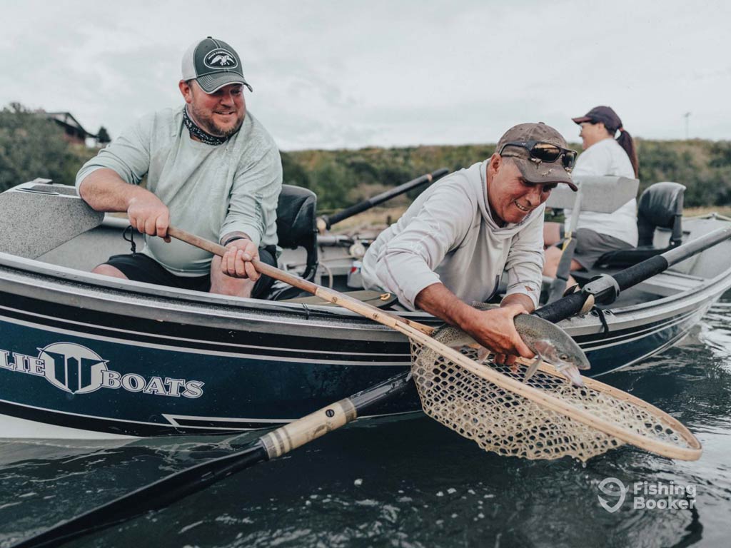 Three people on a boat are fishing in Canada. Two men are holding fishing nets, one with a fish inside, while another person sits in the background. The water and trees create a serene setting.