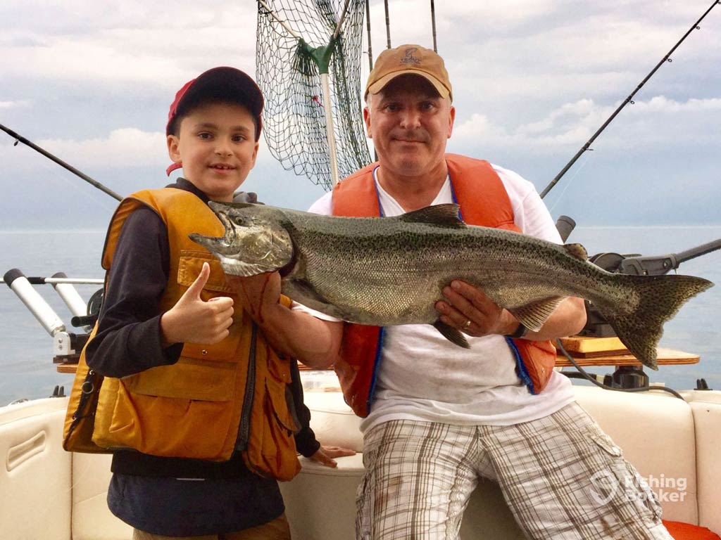 Two people on a boat in Canada are proudly holding a large Salmon. The young angler's giving a thumbs-up, and both are wearing caps and life jackets, capturing the joy of fishing.